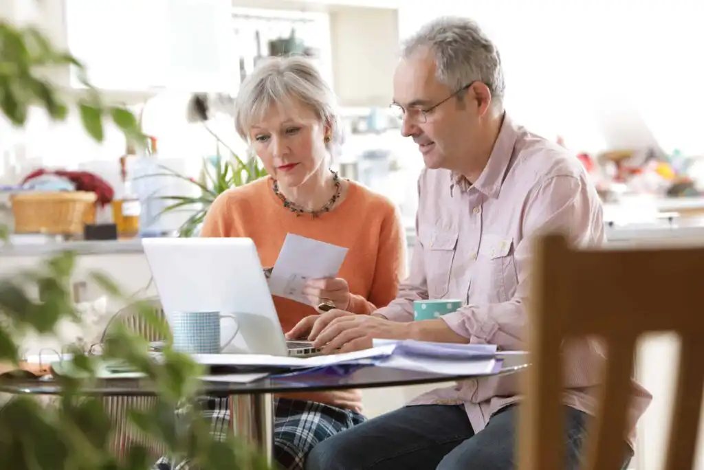 An older woman and man sit at a table in a bright kitchen, looking at documents and a laptop together. Papers and a coffee mug are on the table, suggesting they are working on finances or bills.