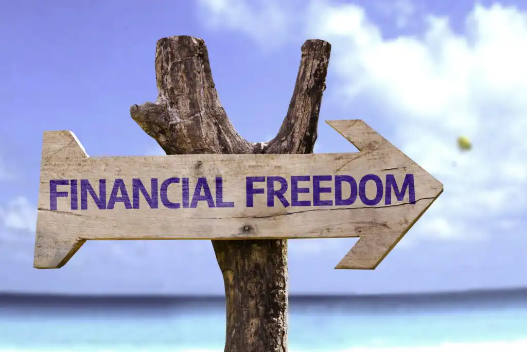 A wooden arrow sign with the words FINANCIAL FREEDOM points to the right. The sign is attached to a tree stump, with a blurred beach and blue sky in the background.