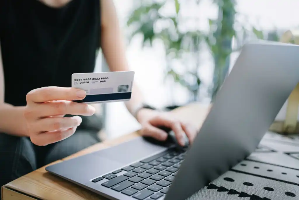 A person holds a credit card in one hand while using the other hand to type on a laptop keyboard, suggesting online shopping or online payment. The background is softly blurred with some greenery visible.