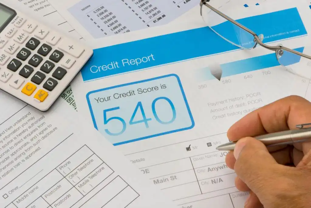 A hand holding a pen fills out forms next to a calculator, glasses, and a credit report showing a credit score of 540. Various paperwork with financial details is spread on the desk.