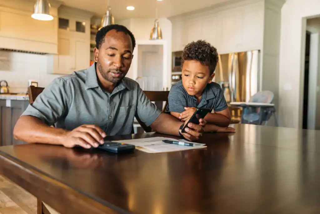 A man sits at a kitchen table using a calculator and holding a phone, while a young boy leans on the table and looks at him, both appearing focused. Papers and pens are spread out on the table.