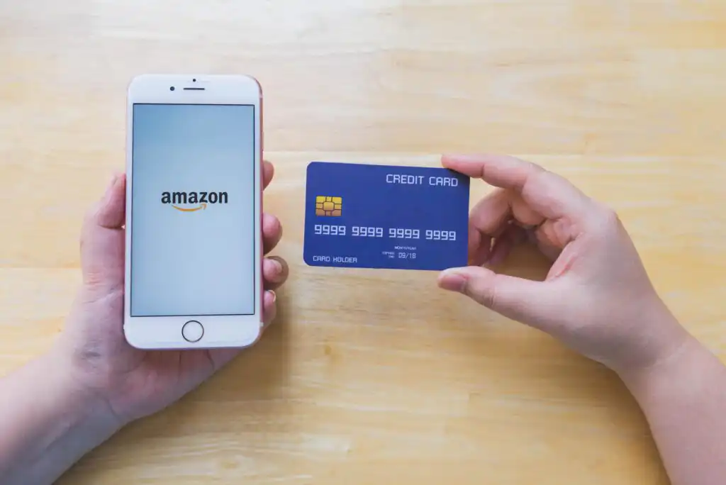 A person holds a smartphone displaying the Amazon logo in one hand and a blue credit card in the other, over a light wooden surface.