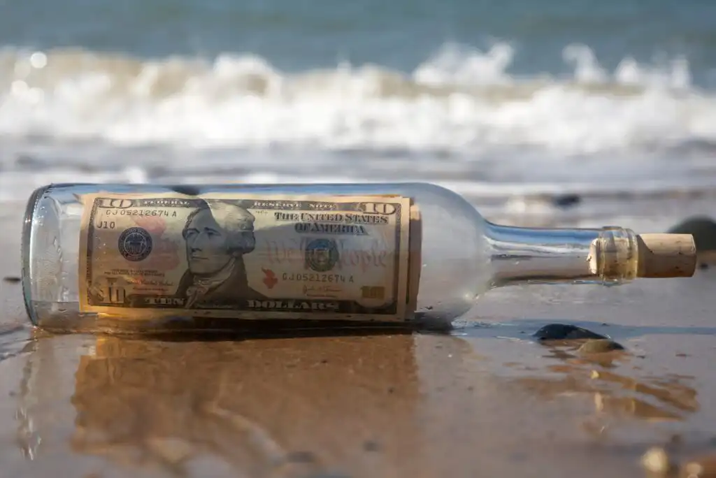 A glass bottle with a cork containing a ten-dollar bill lies on wet sand at the beach, with ocean waves in the background.