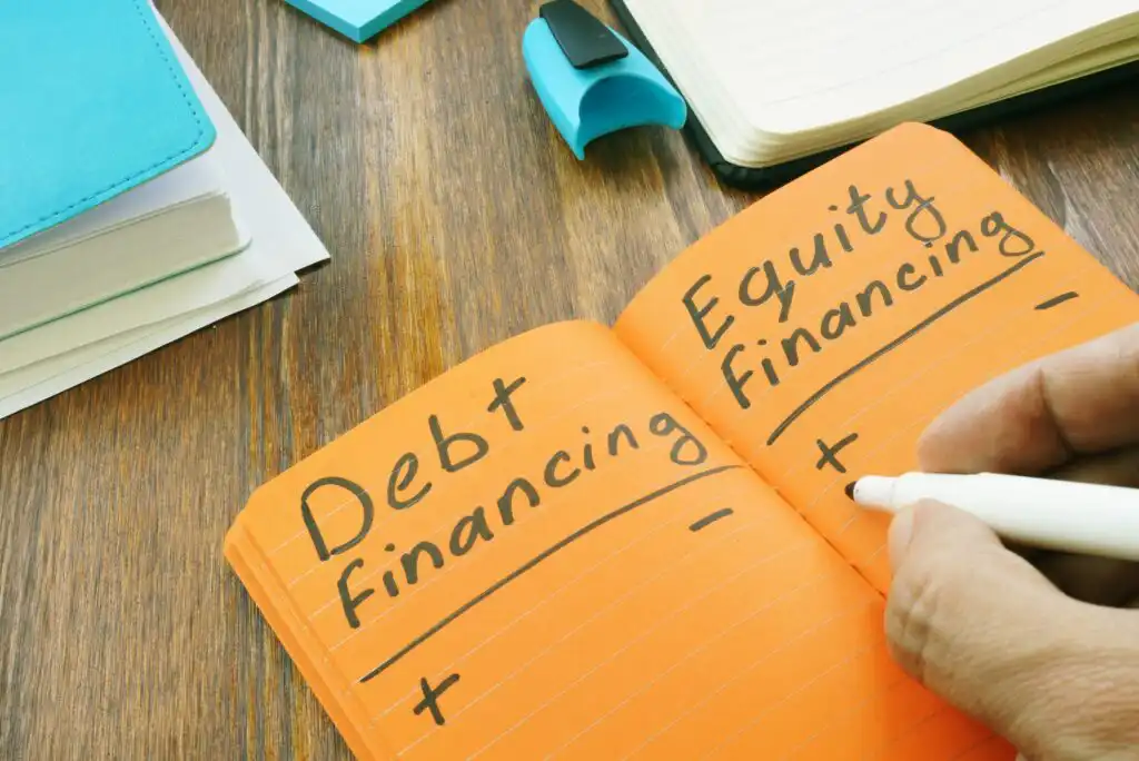 An open orange notebook displays handwritten notes comparing Debt financing and Equity financing with plus and minus signs. A hand writes with a white marker. Office supplies and notebooks are on the wooden desk.