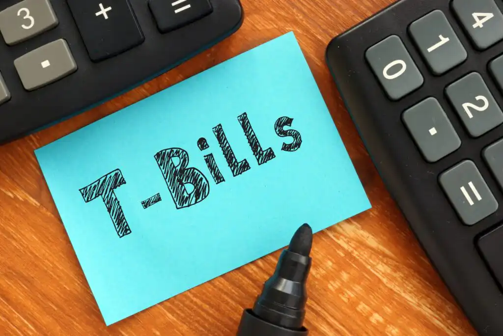 A blue sticky note with “T-Bills” written on it in bold letters is placed on a wooden surface, surrounded by two calculators and a black marker.