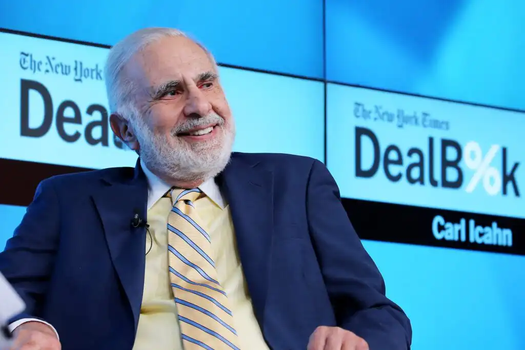An older man in a suit and striped tie smiles while seated in front of a screen displaying “The New York Times DealBook” and “Carl Icahn.”.