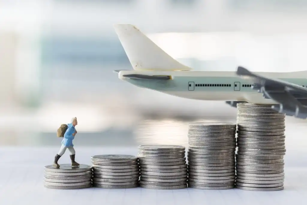 A figurine of a traveler with a backpack stands on stacked coins, leading up to a model airplane, symbolizing the rising costs of travel.