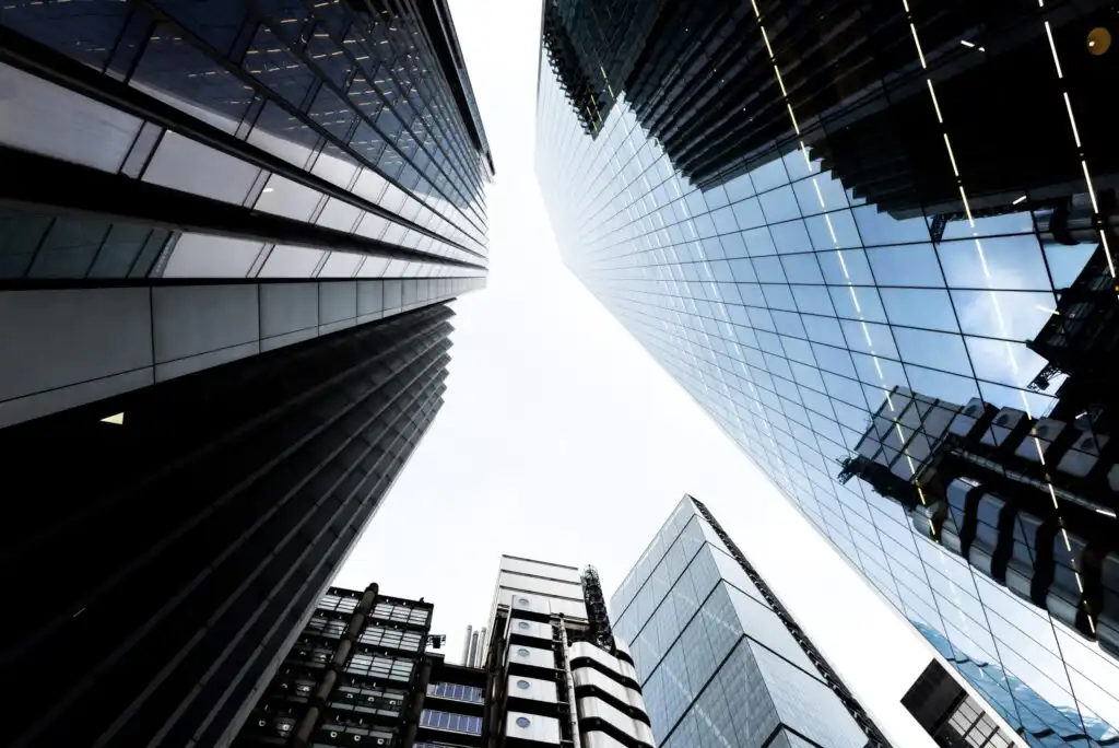 Looking up at several modern skyscrapers with glass and steel facades, reflecting the sky and surrounding buildings, creating a dramatic urban cityscape.