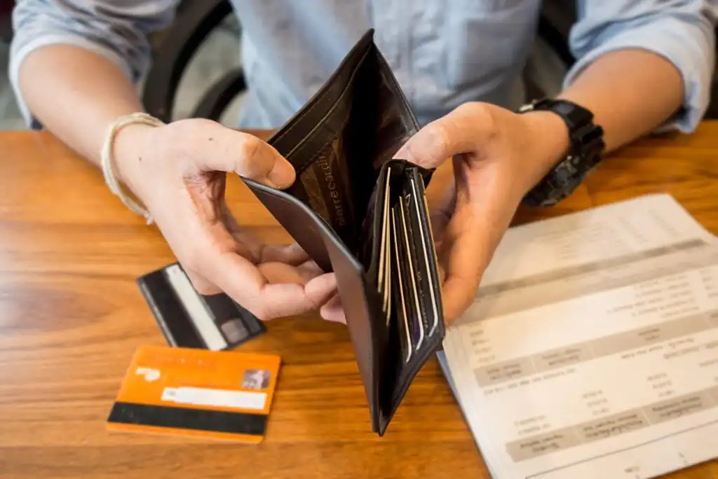 A person holding an open, mostly empty wallet over a table with credit cards and a bill or receipt, wearing a watch and a light blue shirt.