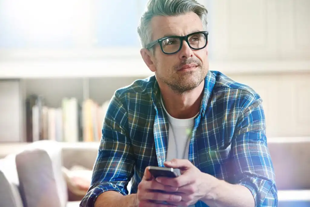 A middle-aged man with gray hair and glasses, wearing a blue plaid shirt, sits indoors holding a smartphone and looking thoughtfully into the distance. Bookshelves and soft sunlight are visible in the background.