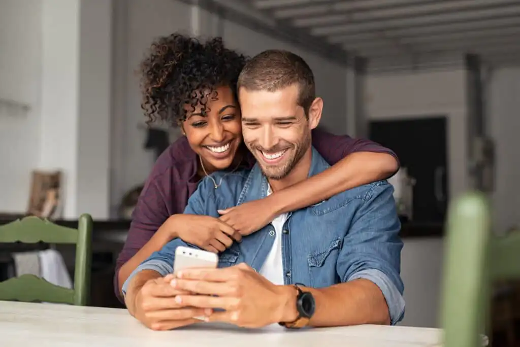 A smiling couple sits at a table, with the woman hugging the man from behind as they look at a smartphone together. They appear happy and relaxed in a bright, modern kitchen setting.