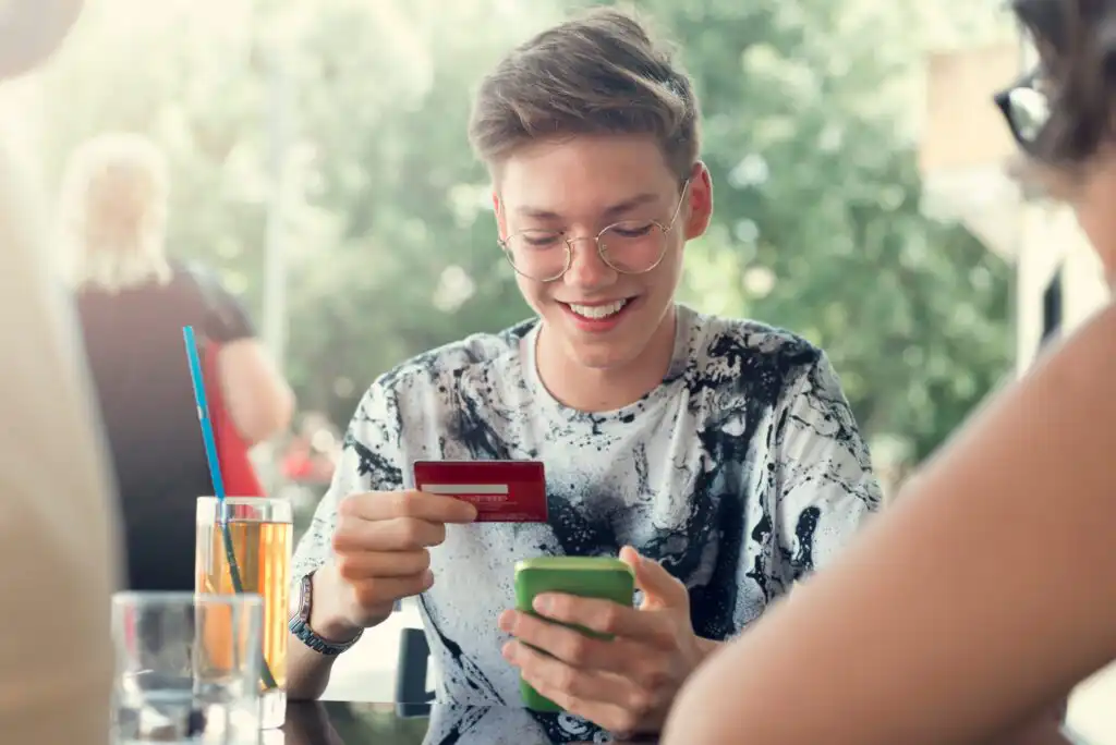 A smiling young person with glasses holds a credit card in one hand and a smartphone in the other, sitting at an outdoor table with drinks, appearing to make an online purchase.