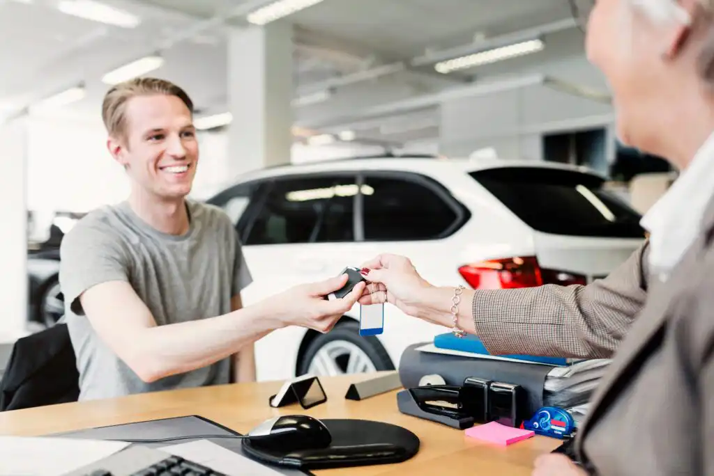 A smiling man receives car keys from another person across a desk in a bright car dealership, with a white car visible in the background.