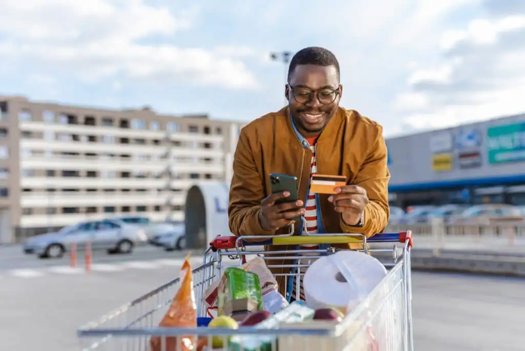 A smiling man stands outside with a full shopping cart, holding a smartphone and a credit card. He appears to be making an online payment or checking something on his phone in a parking lot.