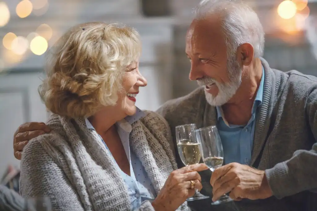 An elderly couple sits close together, smiling warmly at each other while holding and clinking glasses of white wine in a cozy, softly lit setting.