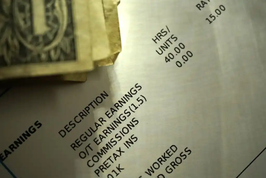 A close-up of a paycheck with details such as regular earnings, overtime, commissions, and hours worked, partially covered by a stack of dollar bills in the upper left corner.