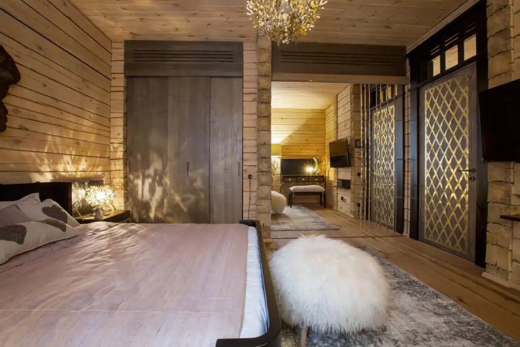 A cozy bedroom with wooden walls and ceiling, a bed with a light pink cover, a fluffy white stool, a chandelier, and mirrored sliding doors reflecting a sitting area in a warmly lit interior.