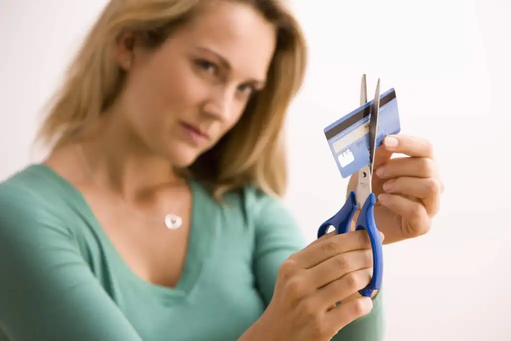 A woman in a green top cuts a blue credit card in half with scissors, suggesting she is canceling or discontinuing its use.