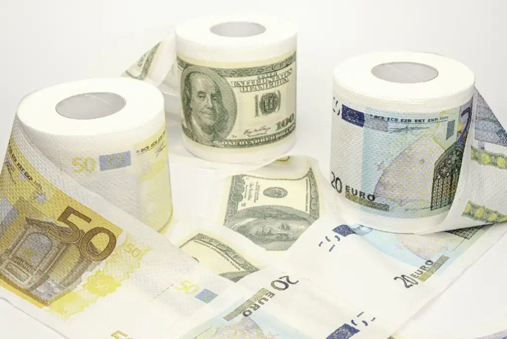 Three toilet paper rolls printed with images of US dollar and Euro bills are displayed, with some sheets unrolled and scattered, alongside real banknotes on a white background.