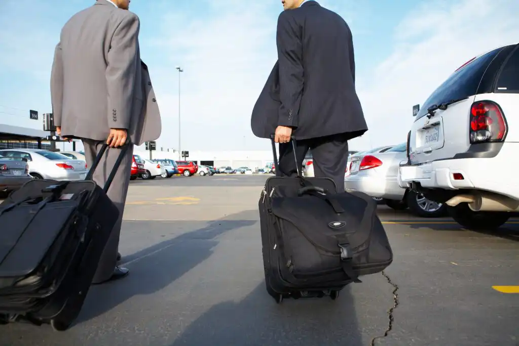 Two people in business suits walk through a parking lot with rolling suitcases on a sunny day, surrounded by parked cars. The photo is taken from behind at a low angle.