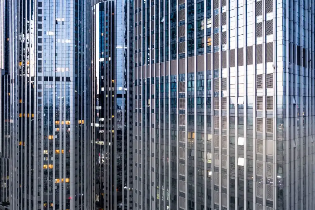 Close-up view of several tall modern skyscrapers with glass facades reflecting light and nearby buildings, showing a grid of windows with some lights on inside.