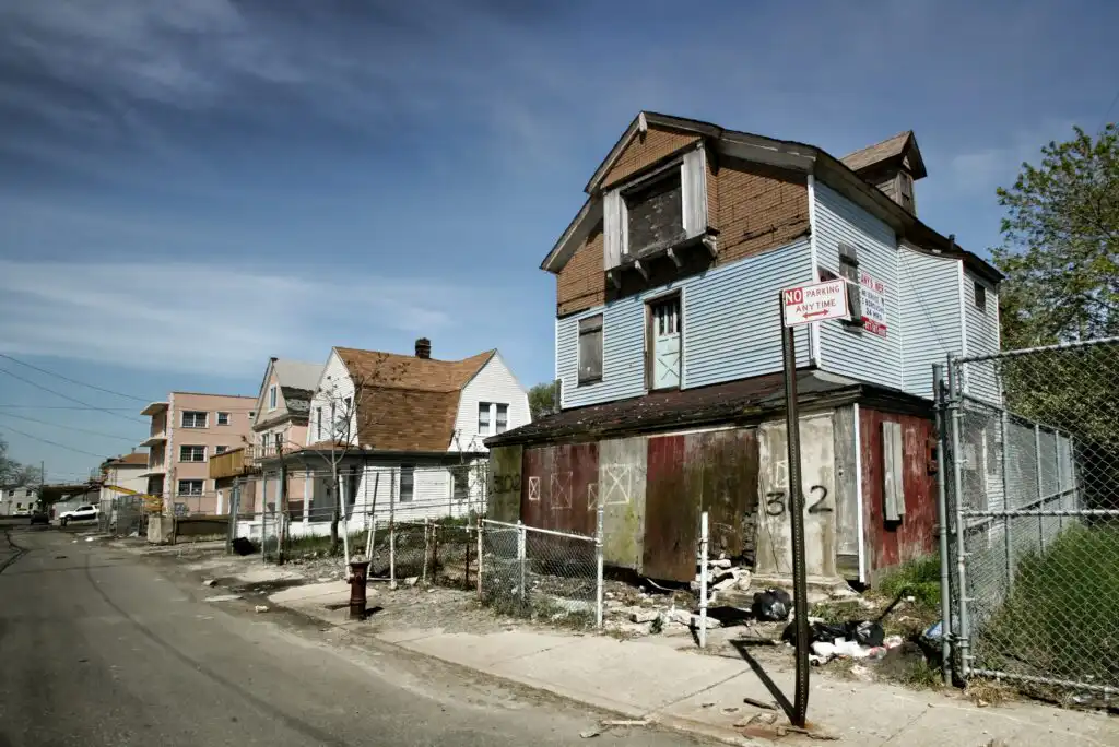A rundown, boarded-up house with peeling paint and debris sits on a neglected street next to a chain-link fence. Nearby houses also appear worn. A no parking sign is visible, under a clear blue sky.