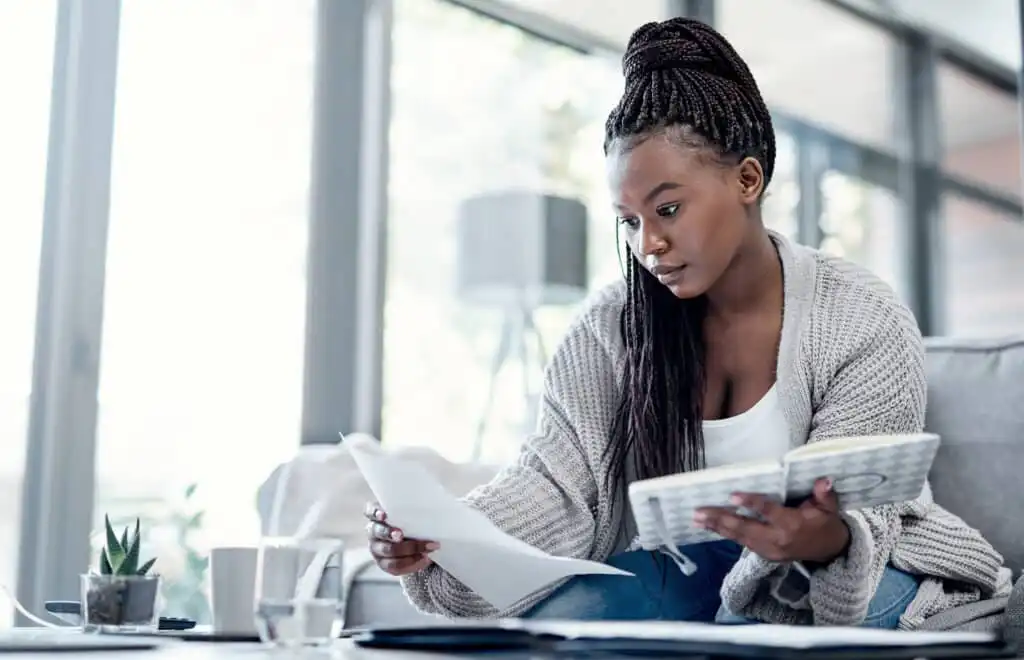 A woman sits on a couch holding papers and a notebook, appearing focused as she works. She wears a cozy sweater and is surrounded by natural light from large windows. A small plant and a glass of water are on the table nearby.
