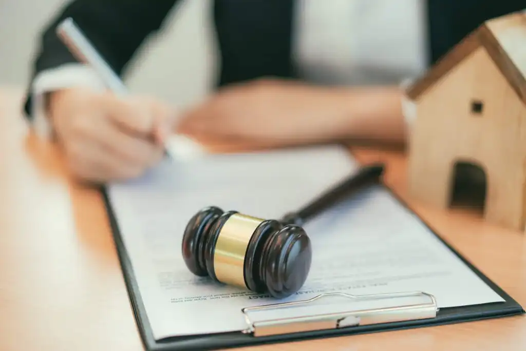 A person signing a document on a clipboard, with a wooden gavel and a small house model on the desk, suggesting a legal or real estate context.