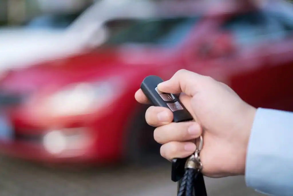 A person’s hand holding a remote car key and pressing a button, with a blurred red car visible in the background.