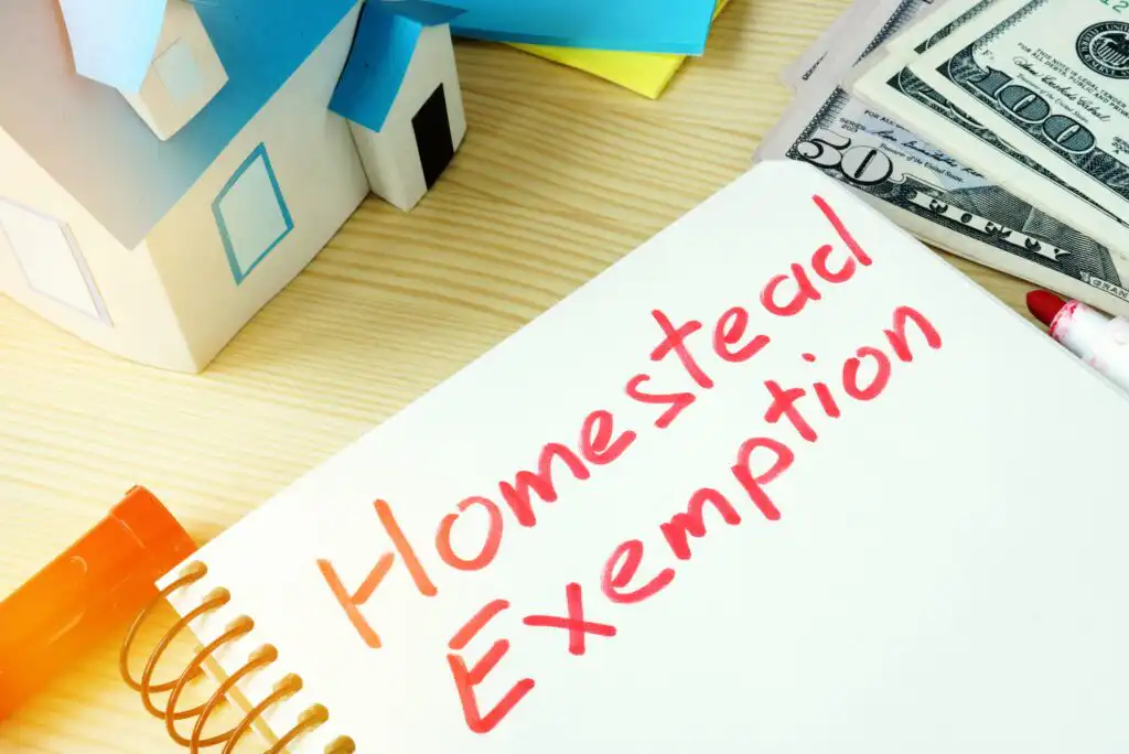 A notebook with “Homestead Exemption” written in red marker sits on a desk beside a model house, a marker, and a stack of dollar bills.