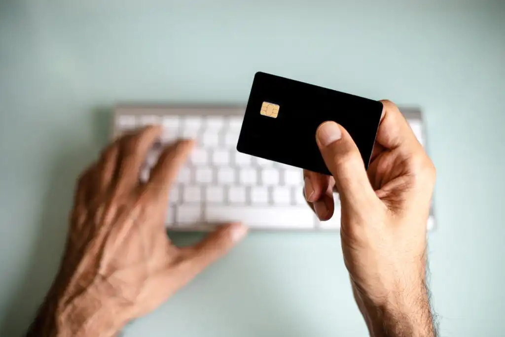 A person types on a white computer keyboard with one hand while holding a black credit card with the other hand, suggesting online shopping or payment. The background is blurred and light-colored.