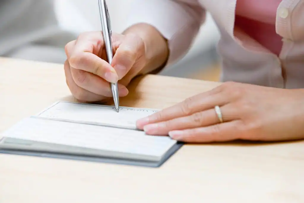 A person wearing a ring writes on a check with a pen at a light-colored wooden desk. The checkbook is open, and only their hands and part of their torso are visible.