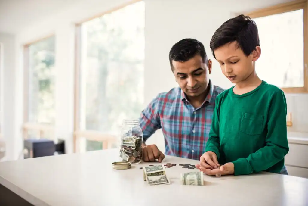 A man and a boy count coins and bills together at a kitchen counter. A jar of money, loose coins, and cash are spread on the white surface. Sunlight streams in through large windows in the background.