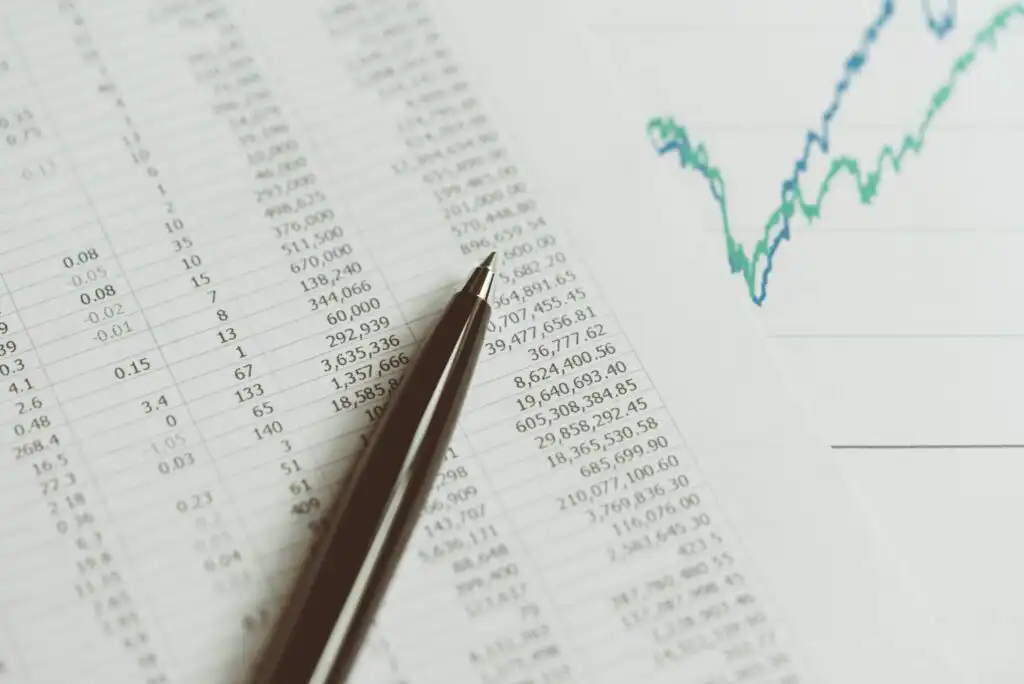 A pen rests on a financial spreadsheet filled with numbers, next to a line graph showing fluctuating data, suggesting financial analysis or business planning.