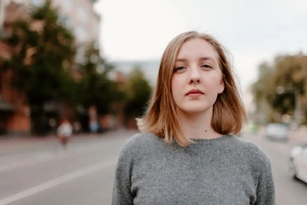 A young woman with straight, shoulder-length blonde hair and a neutral expression stands outdoors on a city street, wearing a grey sweater. The background is blurred, showing buildings and trees.