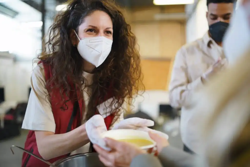 A woman wearing a face mask and gloves hands a bowl of food to another person. She is smiling, and another masked person is visible in the background. The setting appears to be indoors, possibly a community kitchen or food bank.