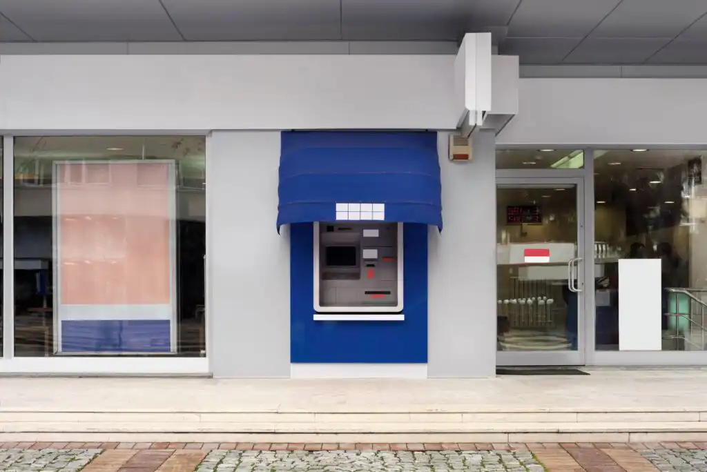 An outdoor ATM with a blue awning is built into the wall of a modern building, next to large glass windows and a glass door. The sidewalk in front is paved with tiles and cobblestones.