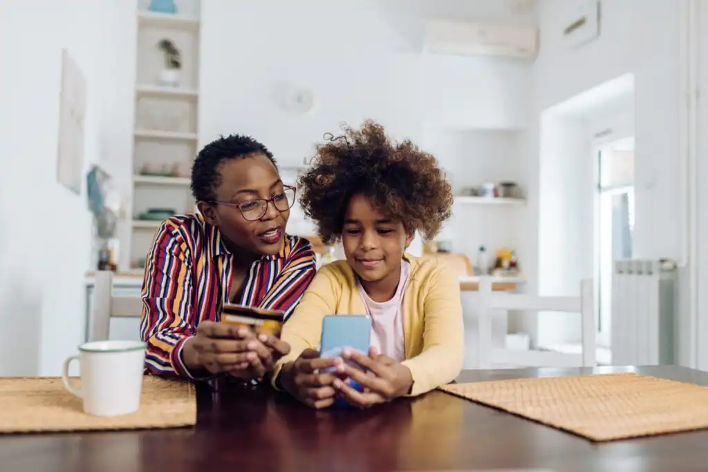 A woman and a young girl sit at a table, smiling and looking at a smartphone together. The woman holds a credit card, suggesting they might be online shopping or learning about technology.