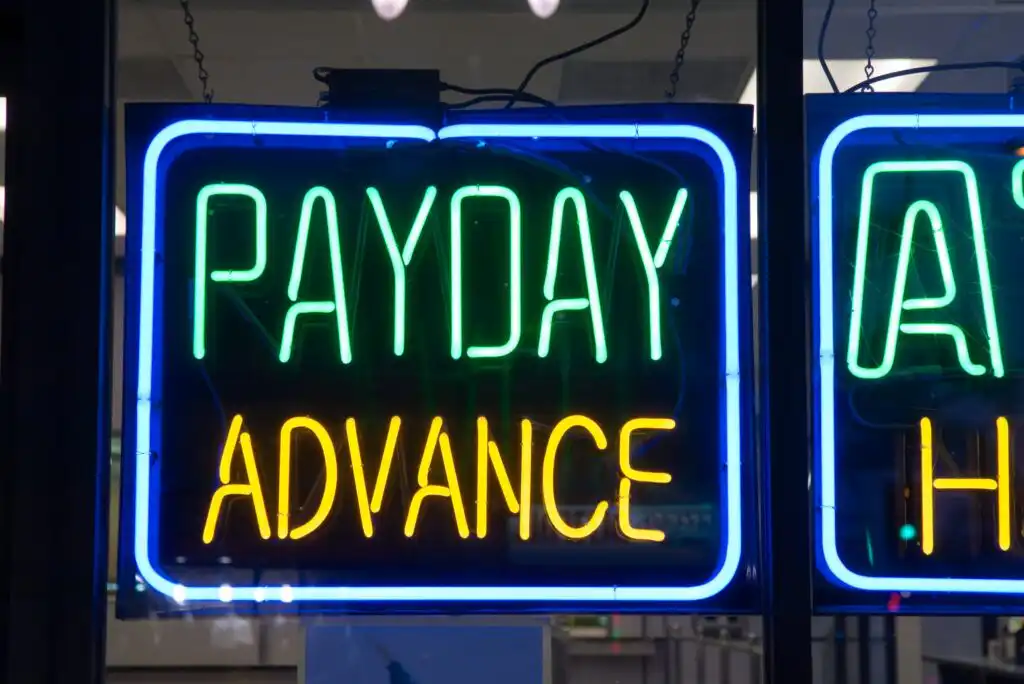 A neon sign in a window displays the words PAYDAY ADVANCE, with PAYDAY in green and ADVANCE in yellow, indicating a business that offers payday loans or cash advances.