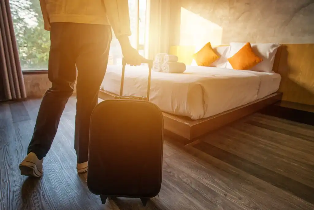 A person with a suitcase enters a sunlit hotel room featuring a neatly made bed with three orange pillows and towels, large windows, and wooden floors.