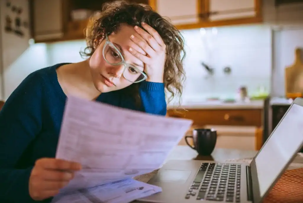 A woman with glasses looks stressed while reviewing papers at a desk with a laptop and a mug, sitting in a kitchen.