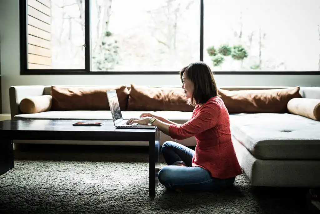 A woman sits cross-legged on the carpet in a living room, working on a laptop at a low coffee table. She is focused on the screen, with a sofa and large window in the background.