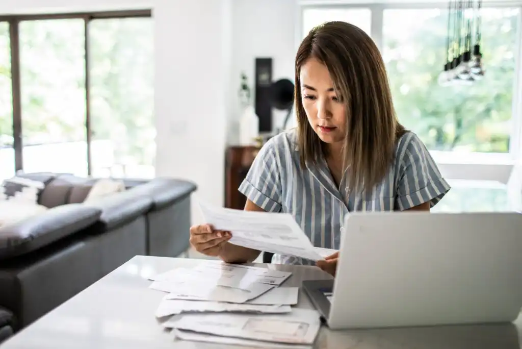 A woman sits at a table in a bright living room, looking at papers while working on a laptop. Several documents are spread out in front of her, and she appears focused and concerned.