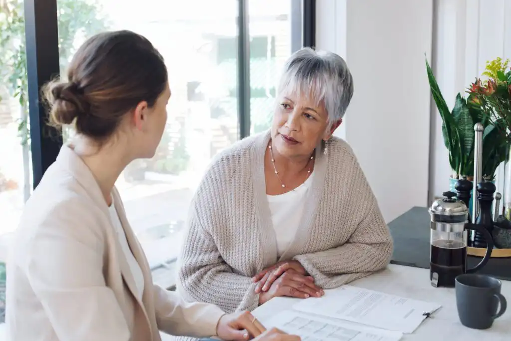 Two women sit at a table talking seriously. One has gray hair and wears a beige sweater, the other has brown hair tied back and wears a light blazer. Papers and a coffee press are on the table. Sunlight comes through a window behind them.