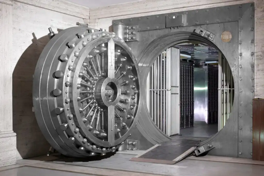 A large, circular metal bank vault door is open, revealing a secure room with metal safety deposit boxes inside. The heavy door features numerous bolts and a complex locking mechanism.