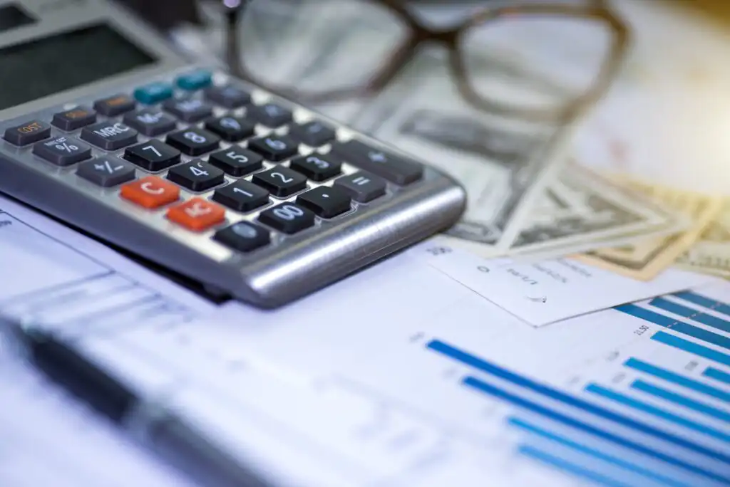 A calculator, eyeglasses, and dollar bills rest on financial documents with charts and graphs, alongside a pen, suggesting a focus on finance, accounting, or budgeting.