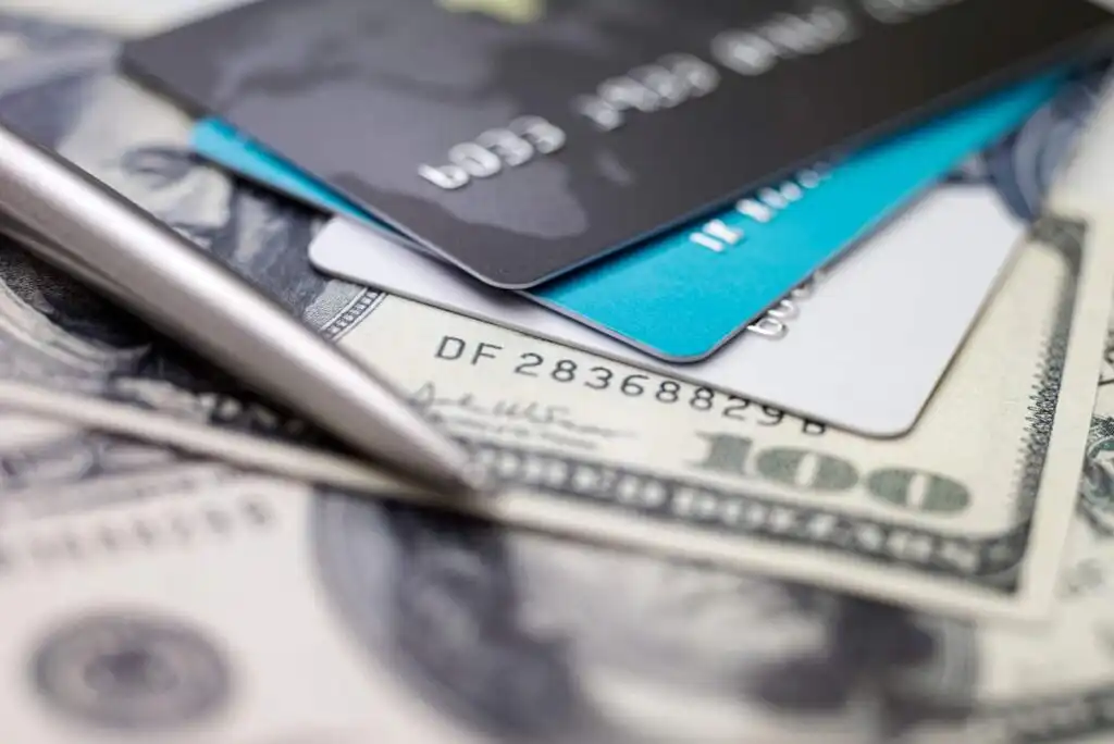 A close-up of several credit cards stacked on top of hundred-dollar bills, with a silver pen lying nearby. The focus highlights the details on the money and cards, symbolizing finance and payments.