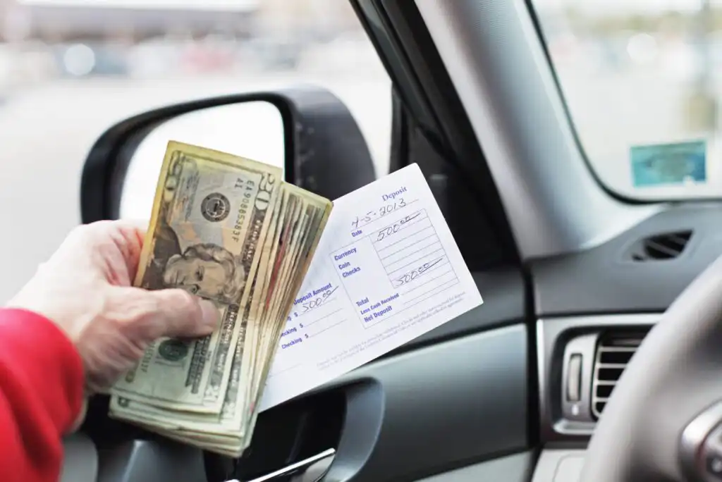 A person in a car holds several US dollar bills and a bank deposit slip next to the driver’s side window, suggesting a bank transaction or deposit.
