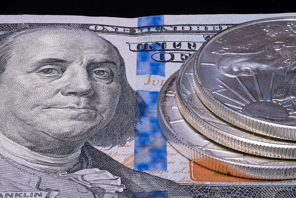 A close-up of a U.S. one hundred dollar bill featuring Benjamin Franklin, with several shiny silver coins stacked on the right side of the bill.