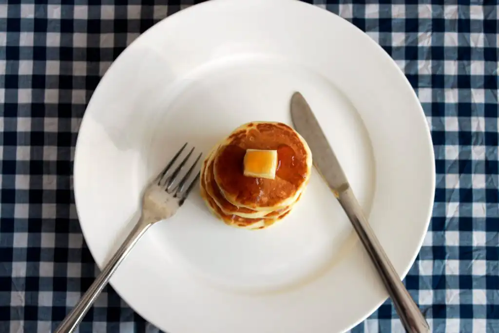 A small stack of pancakes topped with a pat of butter and syrup sits on a white plate, with a fork and knife beside it, set on a blue and white checkered tablecloth.
